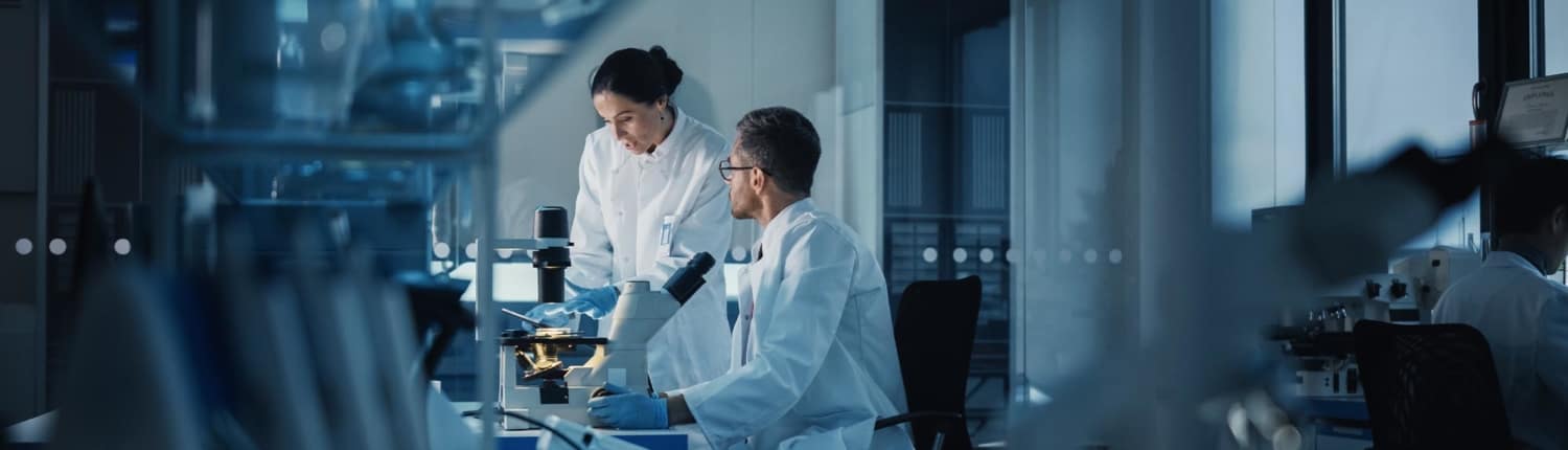 two medical workers assess lab microscope samples in a dimly lit lab